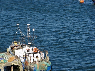A small fishing boat docked at a pier, equipped with various antennas and fishing nets. The surrounding water is a deep blue, and a bright orange buoy floats nearby.