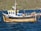 A wooden fishing boat with a blue and white cabin is docked on a body of water, with black tires attached around its perimeter as bumpers. The boat is equipped with various fishing gear, including orange buoys and metal cages. In the background, there is a lush, green hillside.