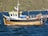 A wooden fishing boat with a blue and white cabin is docked on a body of water, with black tires attached around its perimeter as bumpers. The boat is equipped with various fishing gear, including orange buoys and metal cages. In the background, there is a lush, green hillside.
