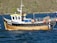 A wooden fishing boat with a blue and white cabin is docked on a body of water, with black tires attached around its perimeter as bumpers. The boat is equipped with various fishing gear, including orange buoys and metal cages. In the background, there is a lush, green hillside.