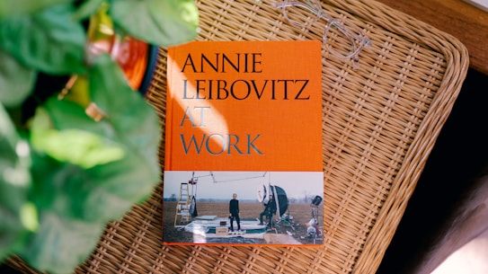A hardcover book titled 'ANNIE LEIBOVITZ AT WORK' is placed on a wicker surface. The book features a bright orange cover with a photograph at the bottom depicting a photography set-up in an outdoor setting. Nearby, there is a pair of eyeglasses and partially visible green potted plants.
