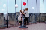 Two children, a girl and a boy, are interacting with a public telescope on an outdoor observation deck. The girl is wearing a colorful skirt and red shoes, while holding a white balloon. The boy is wearing dark pants and a grey shirt and is holding a red balloon. They appear engaged and curious as they look through the telescope. The scene is set against a backdrop of a glass wall, with reflections visible.