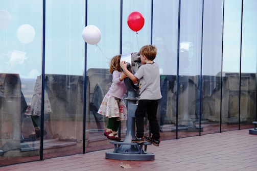 Two children, a girl and a boy, are interacting with a public telescope on an outdoor observation deck. The girl is wearing a colorful skirt and red shoes, while holding a white balloon. The boy is wearing dark pants and a grey shirt and is holding a red balloon. They appear engaged and curious as they look through the telescope. The scene is set against a backdrop of a glass wall, with reflections visible.