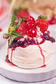 A white meringue dessert topped with a glossy layer of red berry sauce, garnished with fresh strawberries and cherries, presented on a white plate.