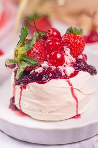 A white meringue dessert topped with a glossy layer of red berry sauce, garnished with fresh strawberries and cherries, presented on a white plate.