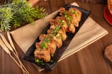 Crispy pieces of fried food, likely pork belly, are arranged neatly on a black rectangular platter. The dish is garnished with chopped green herbs. It is placed on top of a burlap cloth and a wooden surface, with decorative wheat stalks and green plants nearby.
