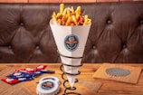 A cone of fries with melted cheese and bacon bits sits in a metal holder on a wooden table. In the background, a leather sofa is visible. To the side, there are several packets of condiments and a plastic container with a sticker that matches the one on the cone.