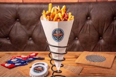 A cone of fries with melted cheese and bacon bits sits in a metal holder on a wooden table. In the background, a leather sofa is visible. To the side, there are several packets of condiments and a plastic container with a sticker that matches the one on the cone.