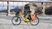 A bright yellow electric bicycle is parked on a rocky riverbank, with a scenic backdrop featuring a large rock formation and a bridge spanning the river. The bicycle is equipped with a basket and orange saddlebags, and the area is surrounded by mountains and trees.