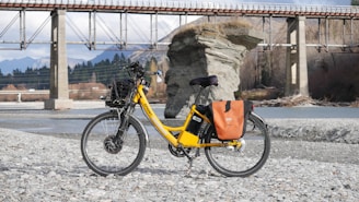 A bright yellow electric bicycle is parked on a rocky riverbank, with a scenic backdrop featuring a large rock formation and a bridge spanning the river. The bicycle is equipped with a basket and orange saddlebags, and the area is surrounded by mountains and trees.