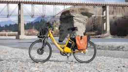 A bright yellow electric bicycle is parked on a rocky riverbank, with a scenic backdrop featuring a large rock formation and a bridge spanning the river. The bicycle is equipped with a basket and orange saddlebags, and the area is surrounded by mountains and trees.