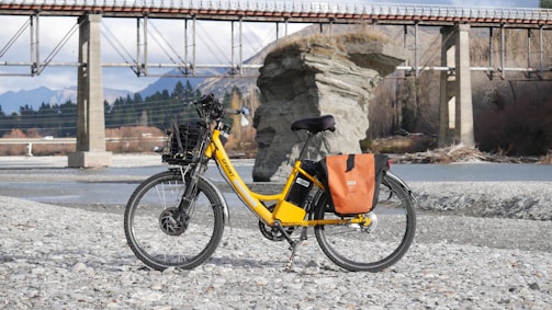 A bright yellow electric bicycle is parked on a rocky riverbank, with a scenic backdrop featuring a large rock formation and a bridge spanning the river. The bicycle is equipped with a basket and orange saddlebags, and the area is surrounded by mountains and trees.