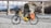A bright yellow electric bicycle is parked on a rocky riverbank, with a scenic backdrop featuring a large rock formation and a bridge spanning the river. The bicycle is equipped with a basket and orange saddlebags, and the area is surrounded by mountains and trees.