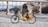 A bright yellow electric bicycle is parked on a rocky riverbank, with a scenic backdrop featuring a large rock formation and a bridge spanning the river. The bicycle is equipped with a basket and orange saddlebags, and the area is surrounded by mountains and trees.
