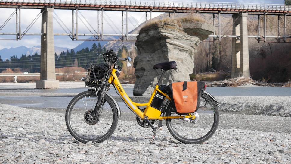 A bright yellow electric bicycle is parked on a rocky riverbank, with a scenic backdrop featuring a large rock formation and a bridge spanning the river. The bicycle is equipped with a basket and orange saddlebags, and the area is surrounded by mountains and trees.