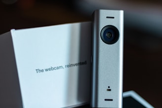 A sleek, modern webcam with a cylindrical design and a metallic finish is positioned next to a white box featuring the text 'The webcam, reinvented'. The background is dark, allowing the product and box to stand out vividly.
