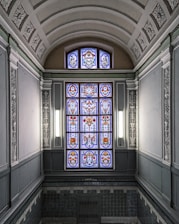 An ornate, stained glass window with intricate floral and geometric patterns is surrounded by decorative plasterwork in a classical architectural setting. The walls and ceiling have detailed relief designs and a soft, neutral color scheme.