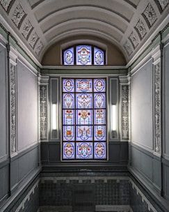 An ornate, stained glass window with intricate floral and geometric patterns is surrounded by decorative plasterwork in a classical architectural setting. The walls and ceiling have detailed relief designs and a soft, neutral color scheme.