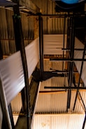 A complex arrangement of industrial pipes and metal structures with sheets of material running through. The setting is dimly lit, creating strong contrasts between light and shadow on the corrugated metal roofing and the structural components.