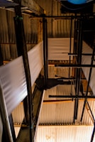 A complex arrangement of industrial pipes and metal structures with sheets of material running through. The setting is dimly lit, creating strong contrasts between light and shadow on the corrugated metal roofing and the structural components.