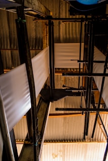 A complex arrangement of industrial pipes and metal structures with sheets of material running through. The setting is dimly lit, creating strong contrasts between light and shadow on the corrugated metal roofing and the structural components.