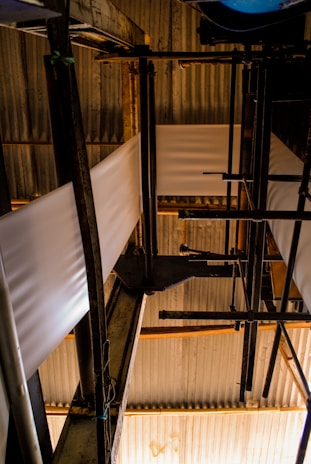 A complex arrangement of industrial pipes and metal structures with sheets of material running through. The setting is dimly lit, creating strong contrasts between light and shadow on the corrugated metal roofing and the structural components.