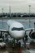 A commercial airplane is parked on a wet airport runway with maintenance and ground crew members in high-visibility vests working around it. In the background, a city skyline can be seen under an overcast sky.