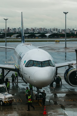 A commercial airplane is parked on a wet airport runway with maintenance and ground crew members in high-visibility vests working around it. In the background, a city skyline can be seen under an overcast sky.