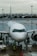A commercial airplane is parked on a wet airport runway with maintenance and ground crew members in high-visibility vests working around it. In the background, a city skyline can be seen under an overcast sky.