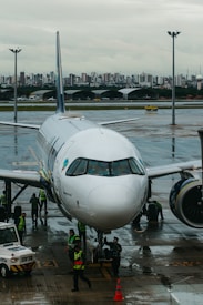 A commercial airplane is parked on a wet airport runway with maintenance and ground crew members in high-visibility vests working around it. In the background, a city skyline can be seen under an overcast sky.