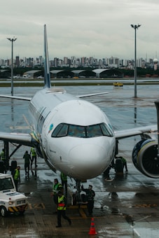 A commercial airplane is parked on a wet airport runway with maintenance and ground crew members in high-visibility vests working around it. In the background, a city skyline can be seen under an overcast sky.