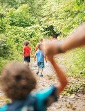 A group of children walk along a narrow, dirt path surrounded by lush greenery. Several kids are dressed in colorful clothing, with vibrant red and blue shirts standing out among the green foliage. The focus is soft, with the foreground showing a child with curly hair reaching out. The setting conveys a sense of adventure and exploration.