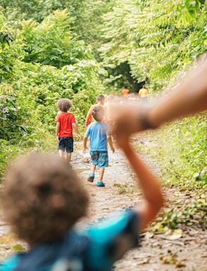 A group of children walk along a narrow, dirt path surrounded by lush greenery. Several kids are dressed in colorful clothing, with vibrant red and blue shirts standing out among the green foliage. The focus is soft, with the foreground showing a child with curly hair reaching out. The setting conveys a sense of adventure and exploration.