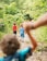 A group of children walk along a narrow, dirt path surrounded by lush greenery. Several kids are dressed in colorful clothing, with vibrant red and blue shirts standing out among the green foliage. The focus is soft, with the foreground showing a child with curly hair reaching out. The setting conveys a sense of adventure and exploration.