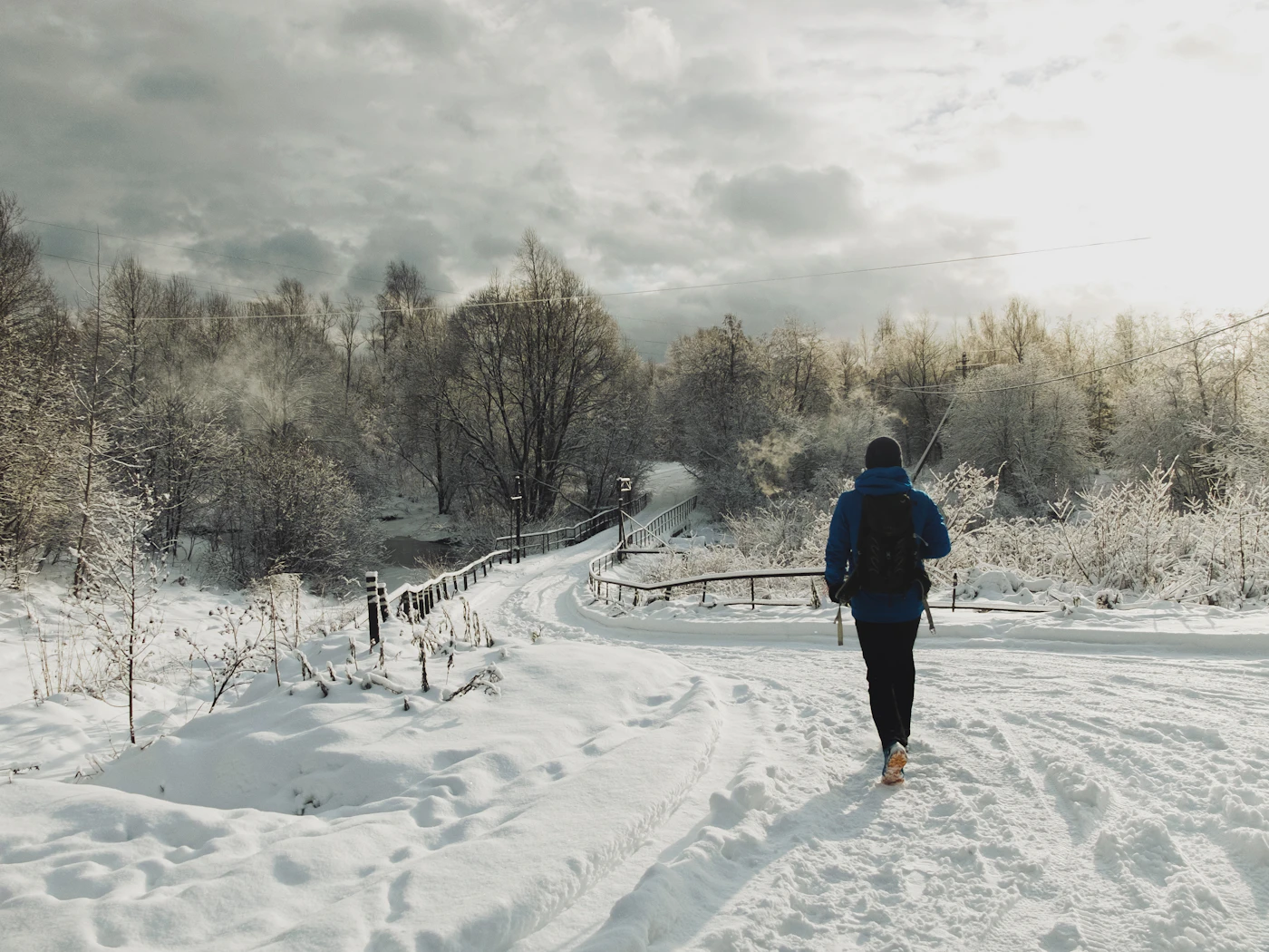 A lone figure with a backpack walking a snowy path toward a small bridge through a quiet Canadian winter landscape