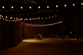A dimly lit outdoor patio area featuring a string of warm white lights hanging above and casting a soft glow. Several tables and chairs are scattered around the space, empty and waiting for occupants. Brick walls and large umbrellas flank the area, creating a cozy but secluded atmosphere.