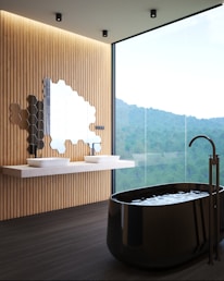 A modern bathroom featuring a large floor-to-ceiling window with a scenic view of trees and mountains. The interior includes a black freestanding bathtub with a sleek faucet, a floating vanity with two white vessel sinks, and a unique cluster of hexagonal mirrors on a wooden panel wall.