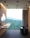 A modern bathroom featuring a sleek black freestanding bathtub filled with water, positioned near a large floor-to-ceiling window overlooking a lush green forest. The room also includes a floating double sink vanity with white basins and black fixtures, mounted on a wooden slat wall. Ceiling lights provide illumination, and a curtain partially covers the window.