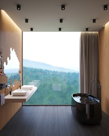 A modern bathroom featuring a sleek black freestanding bathtub filled with water, positioned near a large floor-to-ceiling window overlooking a lush green forest. The room also includes a floating double sink vanity with white basins and black fixtures, mounted on a wooden slat wall. Ceiling lights provide illumination, and a curtain partially covers the window.