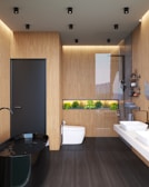 A modern bathroom features minimalistic design elements with wooden panel walls and a sleek black freestanding bathtub. The space includes a white toilet and a contemporary glass shower enclosure. Above the toilet, there is a built-in shelf with green plants providing a touch of nature to the decor. Dual white sinks are installed on a floating counter, and the room is well-lit with recessed ceiling lights.