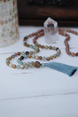 A beaded necklace with a blue tassel is arranged on a white wooden surface. Nearby, there is a clear crystal and a decorative item with a shell-like texture.