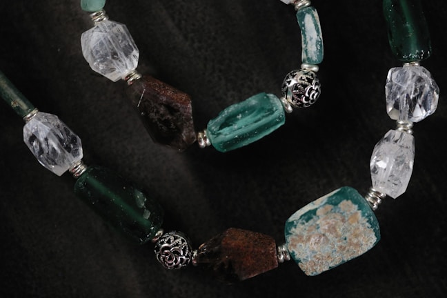 A close-up of a beaded necklace featuring irregularly shaped stones and beads in shades of green, white, and brown. The beads are connected by silver metal connectors with intricate designs.