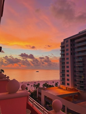 A vibrant sunset paints the sky in shades of pink, orange, and purple over a calm ocean. A yacht is visible on the water, while a sandy beach with scattered palm trees stretches along the shoreline. A large, modern building with balconies overlooks the scene from the right side, framed against the colorful sky.