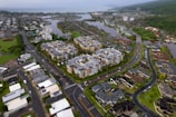 Aerial view of a suburban area with residential buildings and roads. The scene includes large apartment complexes surrounded by smaller houses, lush greenery, and water bodies in the background.