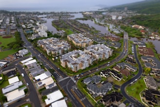 Aerial view of a suburban area with residential buildings and roads. The scene includes large apartment complexes surrounded by smaller houses, lush greenery, and water bodies in the background.