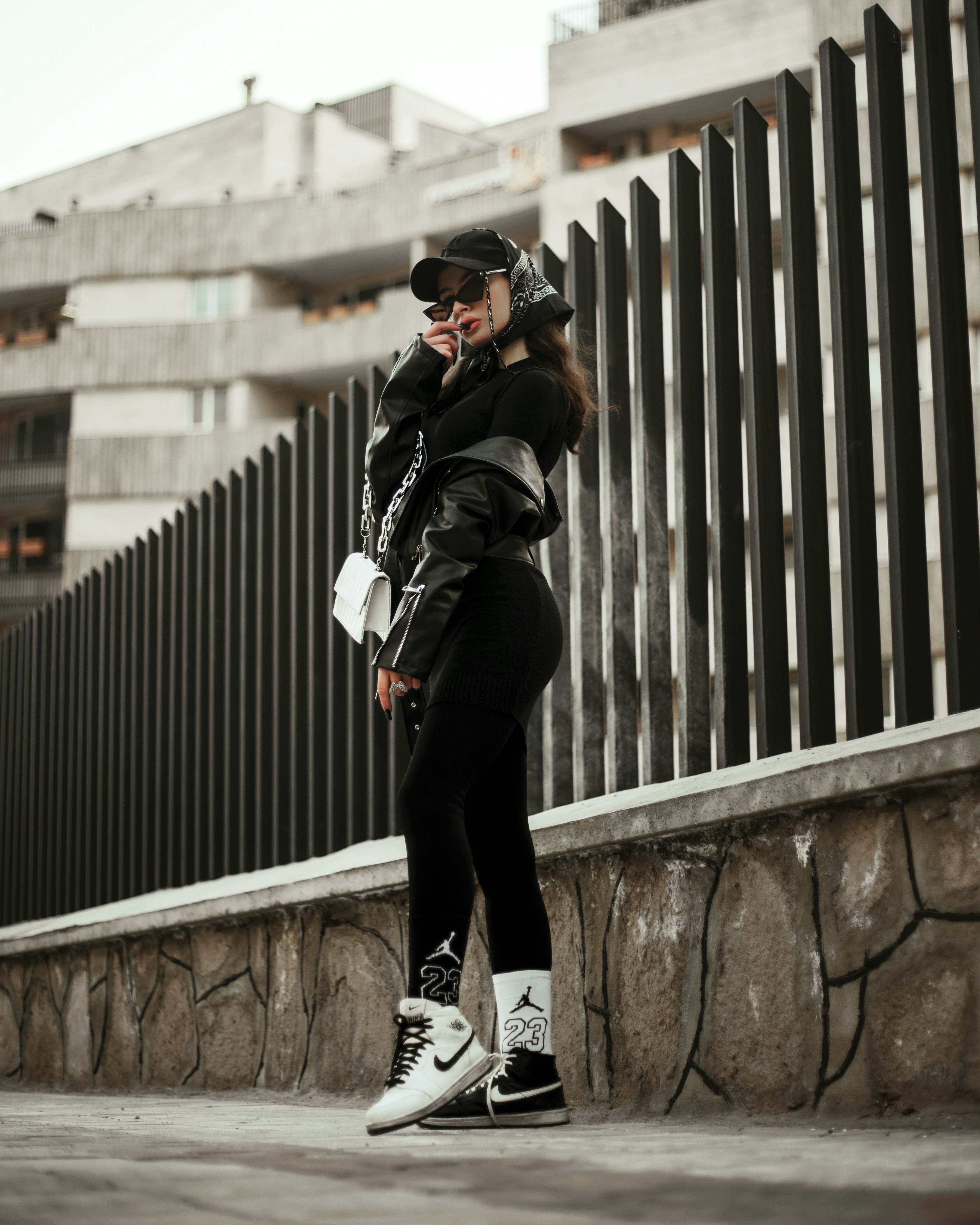 A person wearing urban streetwear poses confidently against a modern architectural background. The outfit includes a black cap, oversized sunglasses, a black leather jacket, and black leggings with white socks and sneakers, along with a white handbag. The setting features a tall, dark fence and a concrete structure in the background, creating a stylish, urban vibe.