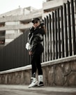A person wearing urban streetwear poses confidently against a modern architectural background. The outfit includes a black cap, oversized sunglasses, a black leather jacket, and black leggings with white socks and sneakers, along with a white handbag. The setting features a tall, dark fence and a concrete structure in the background, creating a stylish, urban vibe.