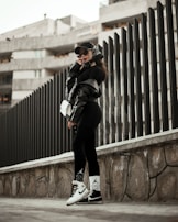 A person wearing urban streetwear poses confidently against a modern architectural background. The outfit includes a black cap, oversized sunglasses, a black leather jacket, and black leggings with white socks and sneakers, along with a white handbag. The setting features a tall, dark fence and a concrete structure in the background, creating a stylish, urban vibe.