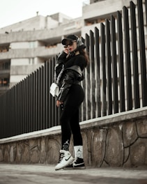 A person wearing urban streetwear poses confidently against a modern architectural background. The outfit includes a black cap, oversized sunglasses, a black leather jacket, and black leggings with white socks and sneakers, along with a white handbag. The setting features a tall, dark fence and a concrete structure in the background, creating a stylish, urban vibe.