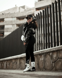 A person wearing urban streetwear poses confidently against a modern architectural background. The outfit includes a black cap, oversized sunglasses, a black leather jacket, and black leggings with white socks and sneakers, along with a white handbag. The setting features a tall, dark fence and a concrete structure in the background, creating a stylish, urban vibe.